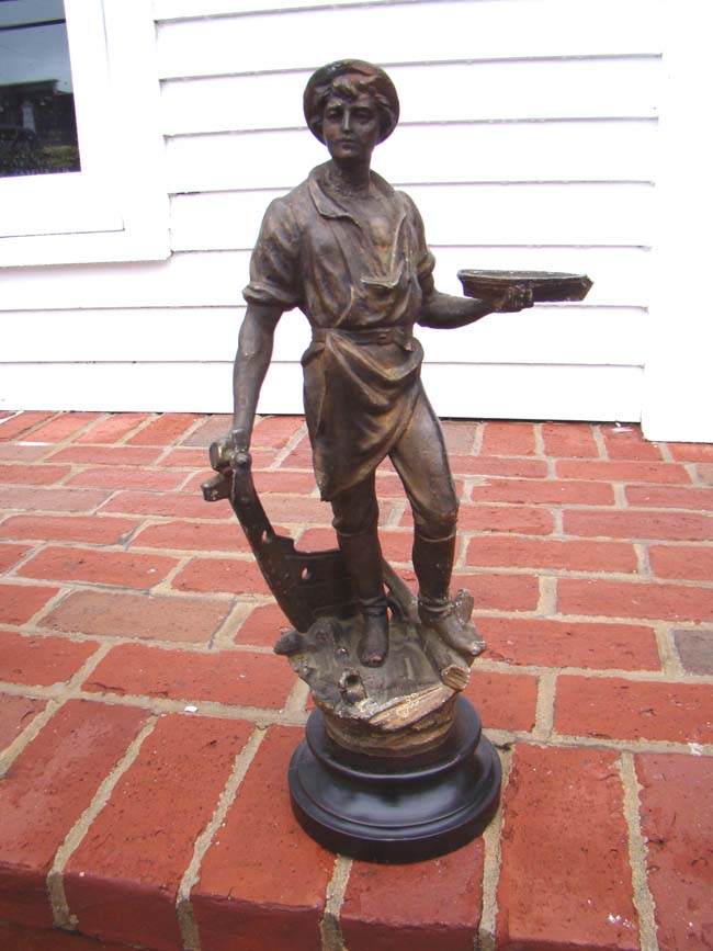 A 19th century metal sculpture of a shipbuilder holding a small ship in one hand and his tool in the other. Circa 1883.